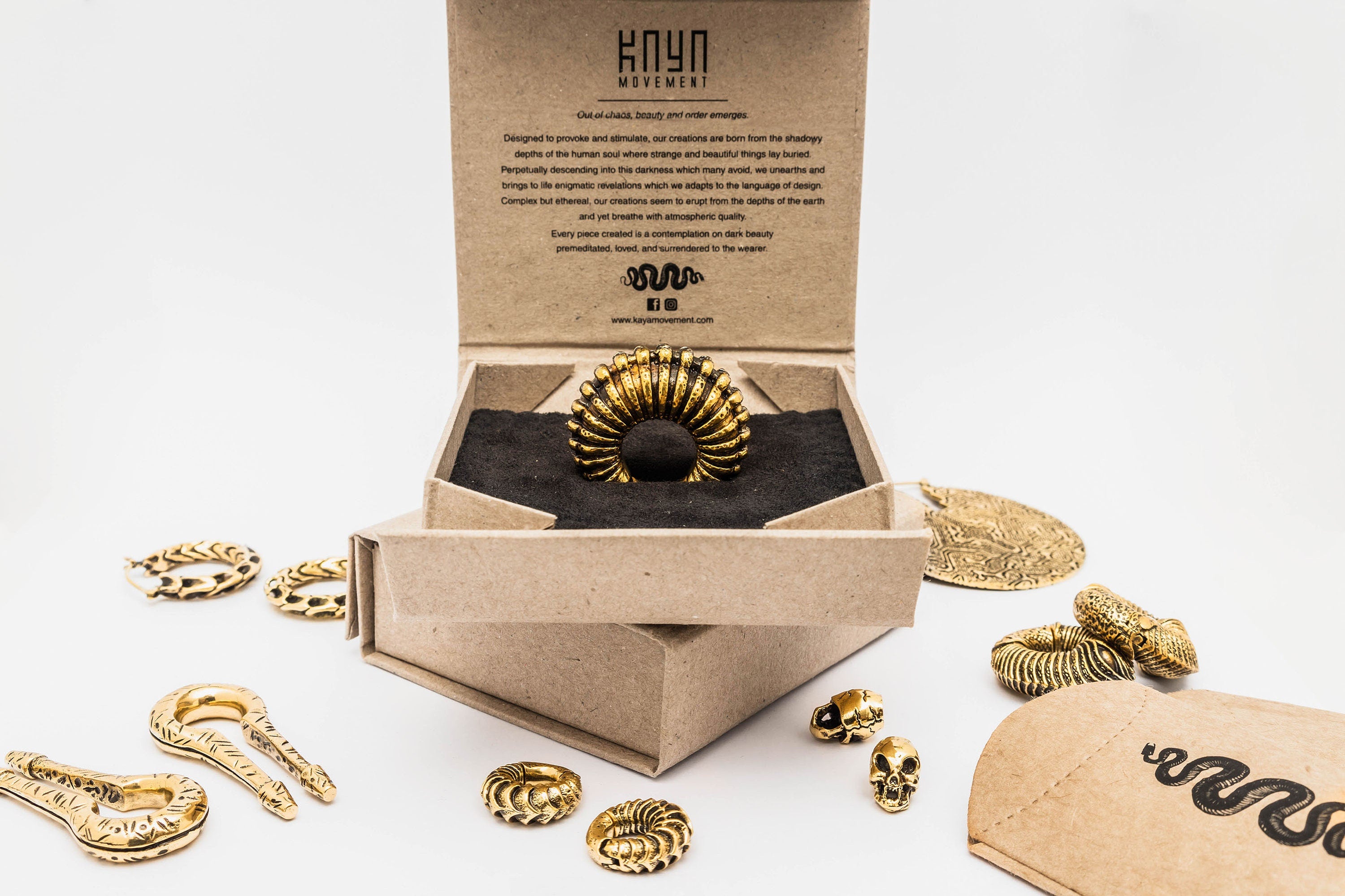 A black and gold ring displayed in an eco-friendly cardboard box exudes a biomechanical-inspired aesthetic. Surrounding the box are various gold jewelry pieces including snake-shaped bracelets, rings, and the Fractal Alien Bio-Organic Circle Ear Weights - Modern Brass 4mm (6ga) Gauges by Kaya Movement. The box lid features text and the Kaya Movement logo. An envelope with a snake print is seen to the right.